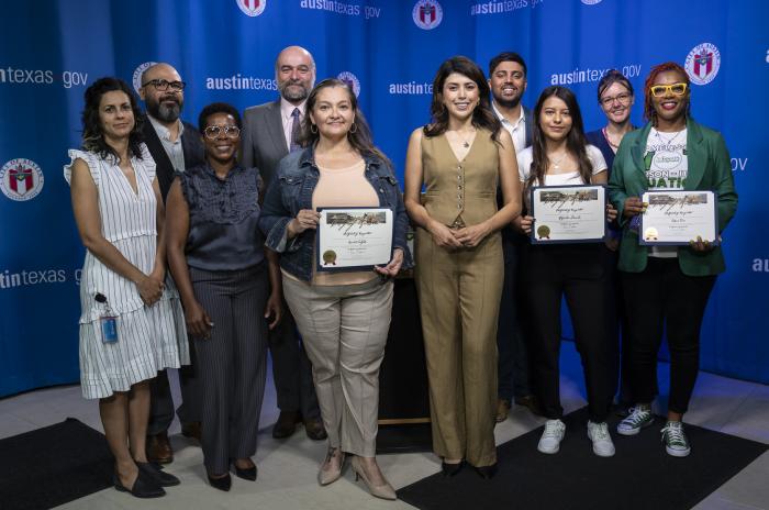 Image shows several Navigators holding certificates in recognition of their community efforts. They are standing with Housing staff and City Council members in front of a blue background with the City of Austin logo and website.