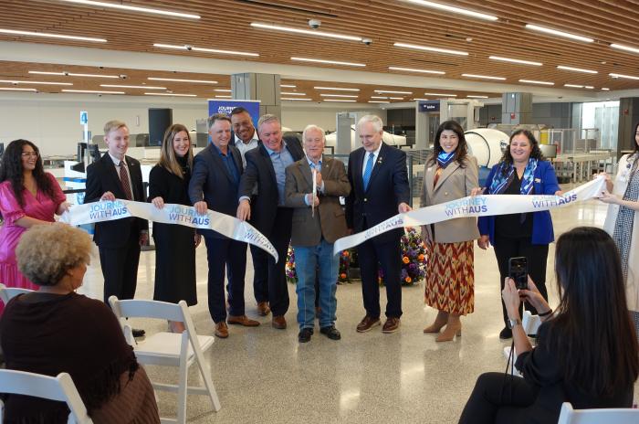 A group of airport and city leaders stand inside a bright, modern terminal area holding a ribbon that reads “Journey With AUS” during a ribbon-cutting ceremony. They smile for photos in front of new security screening equipment, marking the opening of a newly expanded checkpoint space.