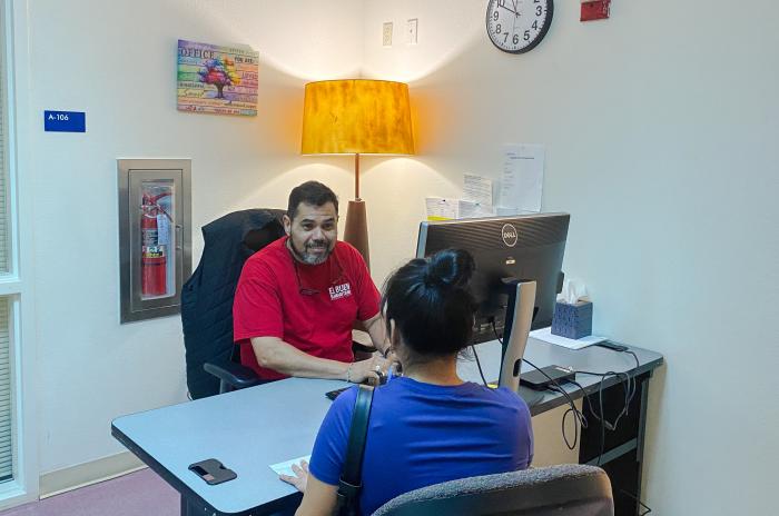 Photo of a male El Buen employee sitting at a desk and assisting a female Austinite, who is sitting on the opposite side of the desk. There is a floor lamp on behind the employee and a clock on the wall.. 