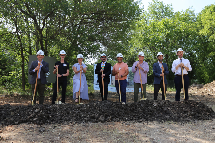 Image shows eight individuals at a groundbreaking. All are standing side-by-side wearing white construction hard hats and holding shovels that are pointed into a mound of dirt. 