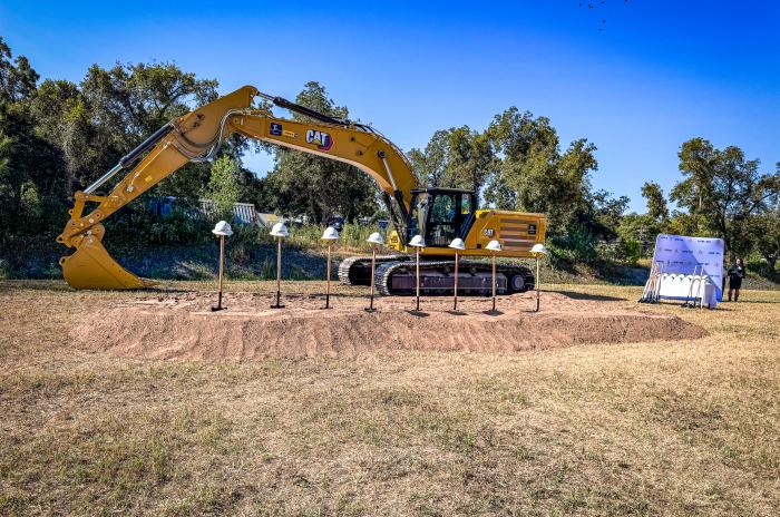 Groundbreaking ceremony setup with a CAT excavator, hard hats on stands, and shovels by a branded backdrop.