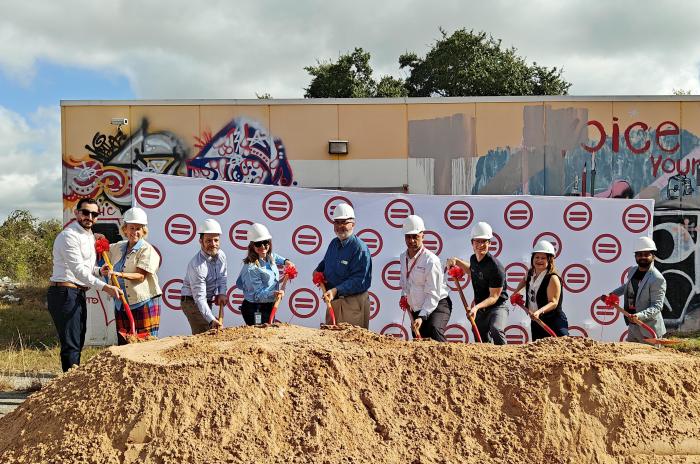 Image shows nine officials, either of which are wearing white hardhats and red holding shovels, standing behind a large mound of dirt. A graffitied trailer is behind them. 