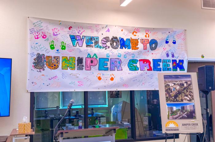 “Welcome to Juniper Creek” banner decorated with colorful handprints and handwritten messages from residents, displayed above a podium and microphone during the Juniper Creek grand opening event. An easel sign shows renderings of the 110-unit affordable housing development.