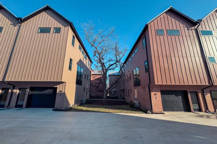 Two modern townhouse buildings face each other across a concrete driveway, with a leafless tree centered between them under a clear blue sky.